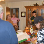 Several people are gathered around a kitchen table with food, snacks, and papers, engaging in conversation in a cozy, homey setting.