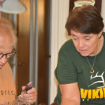 Two women are in a kitchen, one wearing a green Viking shirt, both focused on preparing food together.