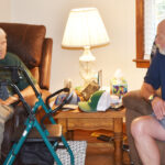 Two older men sit and talk in a cozy living room, with a walker and a lamp on a table between them.