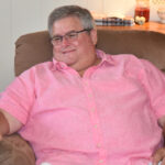 A person wearing glasses and a pink shirt sits comfortably in a brown armchair, smiling.