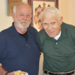Two elderly men standing together indoors, smiling at the camera; one holds a plate of food.