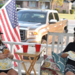 Two people sit on a porch eating, with a USA flag, red cups on a small table, and a white pickup truck parked in the background.