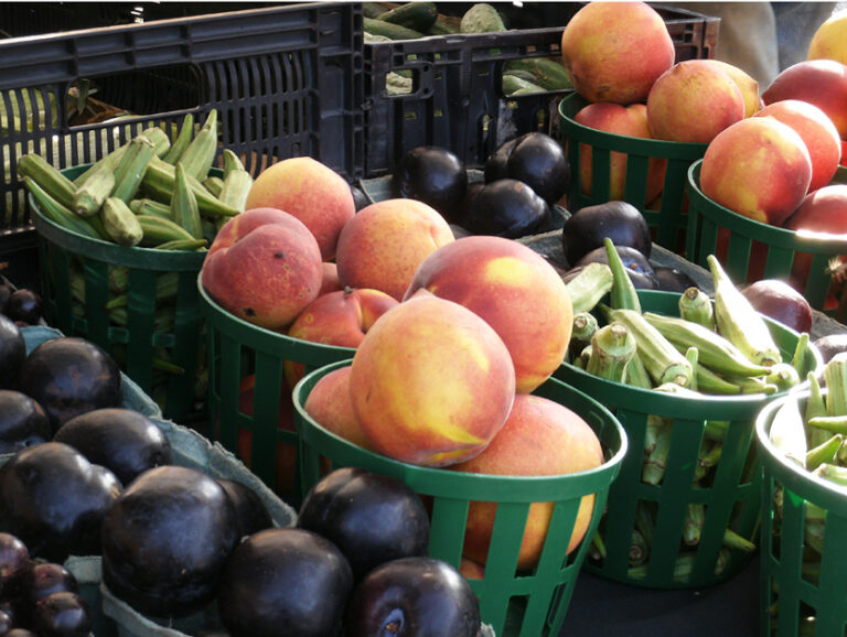Baskets of fresh peaches, plums, and okra are displayed at an outdoor market stand.