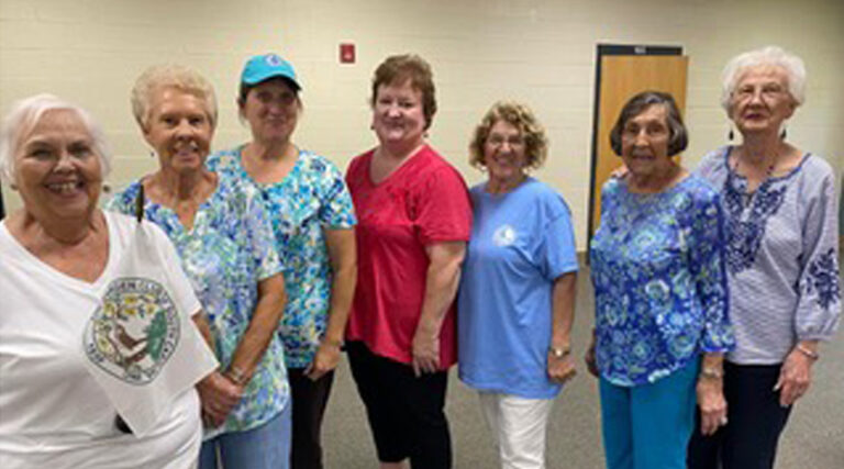 Six women stand in a row indoors, smiling at the camera. They are dressed casually in colorful tops and jeans or pants.