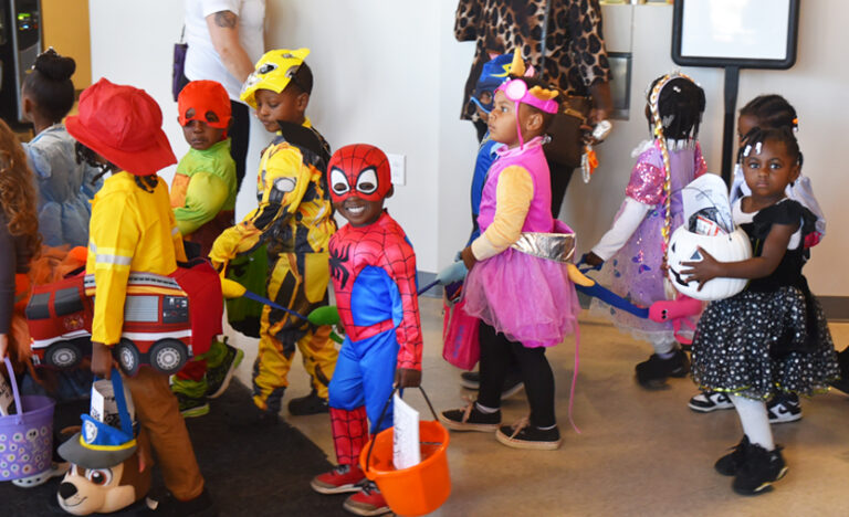 Children dressed in colorful costumes, including superheroes and animals, stand in a line indoors, holding candy buckets and participating in a festive event.