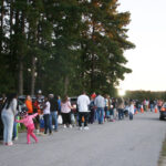 A large group of people, including children in costumes, gather outdoors on a road near trees, possibly for a community or holiday event.
