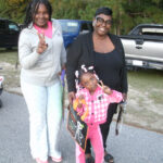Three people standing outdoors, two adults and a child, all smiling and making peace signs. The child holds a colorful bag. Cars and trees in the background.