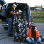 A man arranges Halloween decorations in a hearse while a woman in an orange jacket sits nearby, looking away thoughtfully.
