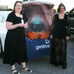 Two women dressed in black stand in front of a Halloween-themed car display with a “gentiva” banner, decorated with lights, outdoors in a parking lot.