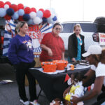 Three women stand behind a decorated table with orange buckets, while a woman and child approach; red, white, and blue balloons are in the background.