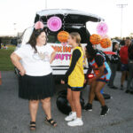 People standing near a decorated car trunk at an outdoor event, with festive hanging decorations and a sign reading “Walk in Mutts.”