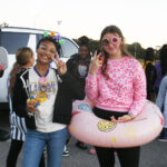 Two young women pose outdoors, smiling and making peace signs, one wearing a pink sweater and inflatable donut, surrounded by other people and vehicles in the background.