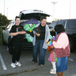 Two women stand by a car decorated with a witch, handing out treats to children in Halloween costumes in a parking lot.
