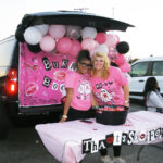 Two women in pink shirts stand by a decorated car trunk with balloons and a table, participating in a festive outdoor event.