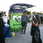 People in costumes stand near a car decorated as a green monster face, with bubbles floating around during a Halloween trunk-or-treat event.
