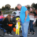 Adults and children, including a child in a yellow costume, gather outdoors. One woman sits handing out candy while others stand in line, enjoying a festive event.