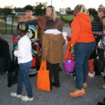 People in costumes gather outdoors, carrying colorful bags, likely participating in a Halloween or fall event.