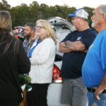 A group of people stand outdoors near a car with an open trunk, casually talking; trees and vehicles are in the background.