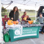 People at a decorated booth with pumpkins and a spider web, under a tent. The tablecloth reads "Anderson Brothers Bank." Outdoor event in daytime.