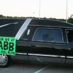 A black hearse with a green sign on the side saying, "Join us at ABB, feel alive," parked outdoors.