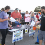 People gather around a table outdoors, handing out papers and treats, while children and adults interact near parked cars during a community event.