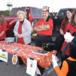 Four adults stand behind a decorated table at a trunk-or-treat event, with a person in a Goofy costume interacting in the foreground. Pickup trucks are in the background.