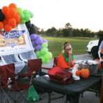 Two people sit at a decorated outdoor table with colorful balloons, pumpkins, and chairs. A banner and open field are visible in the background.
