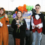 Three people in Halloween costumes stand outdoors, with colorful balloons and a decorated table behind them, holding snacks or treats.
