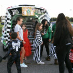 Children in Halloween costumes gather around a decorated car trunk at a trunk-or-treat event in a parking lot during daylight.