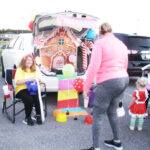 A decorated car trunk with a gingerbread house theme, people standing nearby, one child in costume, and colorful props in a parking lot.