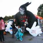 People gather around a large inflatable Grim Reaper decoration at a Halloween event; a child in a blue costume walks in front.