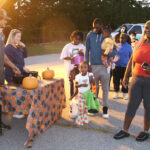 People gather outdoors around a decorated table with pumpkins at sunset. Some children are holding treat bags, suggesting a festive Halloween or autumn event.