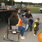 Three adults sit in chairs near a truck, talking and smiling, while a young child in a dinosaur costume stands nearby, holding a pumpkin bucket.