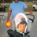 Man in a blue shirt stands behind a stroller holding a pumpkin bucket; a baby in costume sits in the stroller outdoors.