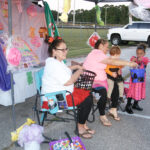 Two women sit at a decorated booth handing out treats to costumed children during an outdoor event, with a white truck and sports field in the background.