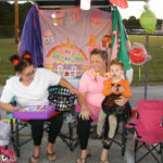 Three people, including a child in a Halloween costume, sit on folding chairs outdoors with colorful decorations and a painted backdrop behind them.