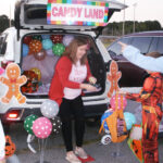 A woman gives out candy from a decorated car trunk with a Candy Land sign, balloons, and gingerbread cutouts at an outdoor event.