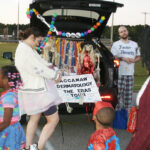 Adults and children in costumes gather by a decorated car trunk with hanging skeletons and a sign, outdoors during daylight, likely at a Halloween or trunk-or-treat event.