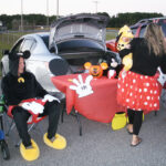 Two people in Mickey and Minnie Mouse costumes set up a decorated table at the back of a car in a parking lot.