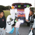 Children in costumes gather around a decorated car trunk with Halloween decorations and inflatable ghosts during a Halloween event outdoors.