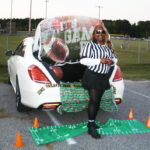 A woman dressed as a referee stands by a decorated car trunk with a football theme, including football props and cones, in an outdoor parking lot.