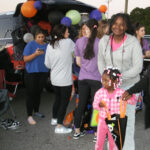 A group of people gathers near a decorated car trunk with balloons; a smiling woman and young girl hold bags in the foreground.