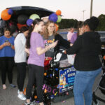 A group of people gather around a decorated car trunk with balloons, participating in a community event outside during the day.