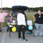 Four people stand near a decorated car trunk with balloons and a table at an outdoor event, possibly a fair or community gathering.