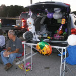 A man sits by a car decorated with Halloween balloons and pumpkins, participating in a trunk-or-treat event in a parking lot.