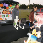 People at a Halloween trunk-or-treat event, with decorations including balloons, a skeleton, and a sign. Children in costumes are holding treat bags.
