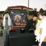 People at a trunk-or-treat event with a decorated car, Halloween backdrop, witch figure, and candy bowls; one person holds a baby in costume.