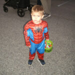 Young boy dressed in a Spider-Man costume holding a green pumpkin bucket, standing on pavement with a stroller and other people in the background.