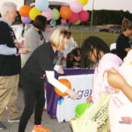 People at an outdoor event with colorful balloons, a woman handing something to a girl near a booth, and others standing nearby.