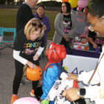A woman hands out candy to children in costumes at an outdoor event, with balloons and people gathered around a table in the background.